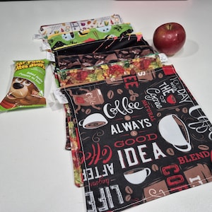 May include: A collection of reusable snack bags with diverse patterns, including coffee-themed and floral designs. A red apple and a snack packet are placed beside the bags. The bags are made of fabric and are designed for carrying food items.