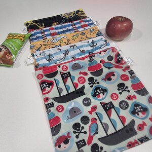 May include: Several reusable snack bags with nautical patterns, including anchors, pirate ships, and lighthouses. A red apple and a snack bar are also present. The bags are made of fabric and are designed for carrying food items.
