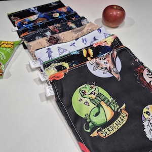 May include: A selection of reusable snack bags with diverse patterns, including a snake with the text "SERPENEARD". A red apple and a snack packet are also visible.