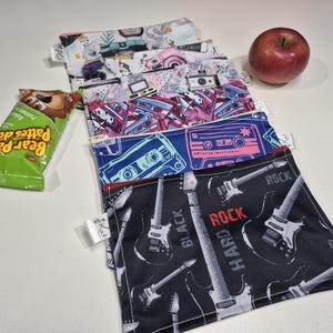 May include: A collection of reusable snack bags featuring designs such as cameras, cassette tapes, and guitars. A red apple and a Bear Paw fruit snack are also visible. The bags are made of fabric and have zippers.