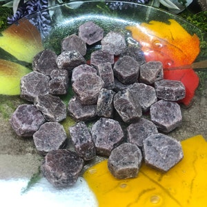 May include: A collection of small, dark purple, faceted ruby crystals in a glass dish. The crystals are arranged in a random pattern and are surrounded by a yellow and orange background.
