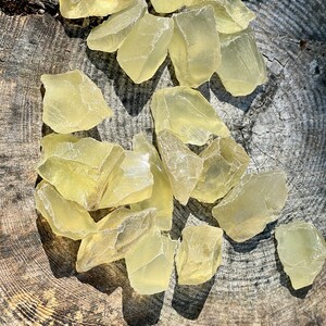 May include: A collection of translucent yellow selenite crystal pieces on a wooden surface. The crystals are various shapes and sizes, with some having a more jagged edge.