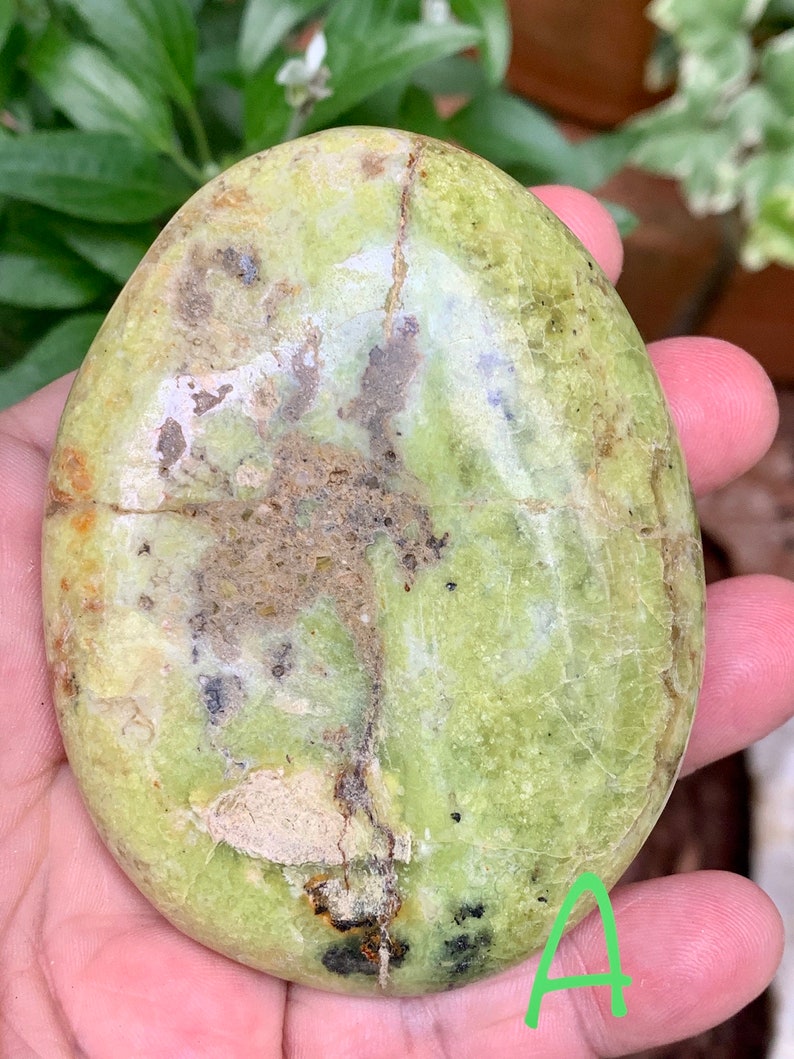 May include: An oval-shaped, polished stone in shades of light green, with brown and darker green patterns. The stone's surface shows natural cracks and imperfections. The letter "A" is written in green on the bottom right corner of the stone.