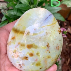 May include: A polished, oval-shaped stone displaying a blend of pale green, white, and brown tones. The stone's surface is smooth, featuring natural patterns and minor cracks. The letter "B" is written in green on the top right.