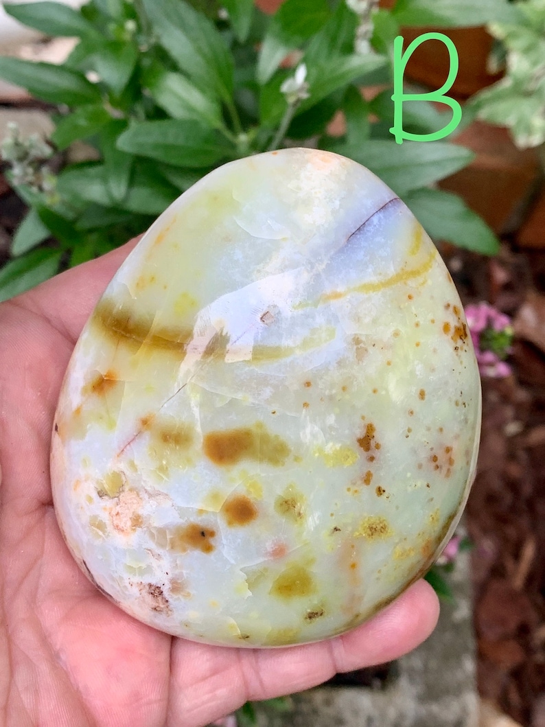May include: A polished, oval-shaped stone displaying a blend of pale green, white, and brown tones. The stone's surface is smooth, featuring natural patterns and minor cracks. The letter "B" is written in green on the top right.