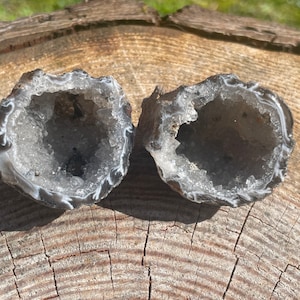 May include: Two grey agate geode halves with white crystal formations inside. The geode halves are sitting on a wooden surface.