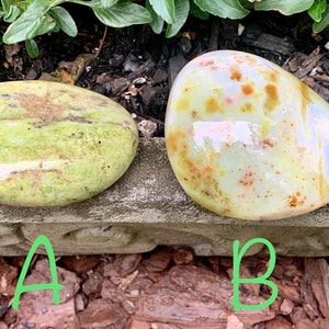 May include: Two polished stones, one oval and the other rounded, are displayed on a concrete surface. The oval stone is pale green with brown markings. The rounded stone is creamy yellow with brown and blue accents. The stones are displayed outdoors.
