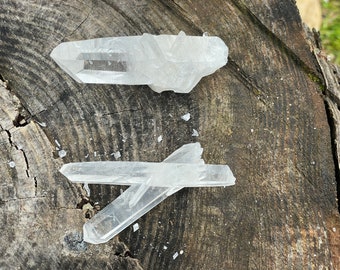 Op de afbeelding: Twee heldere kwartskristallen op een ruw houten oppervlak. De kristallen zijn puntig en hebben een natuurlijke, ruwe uitstraling.