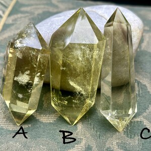 May include: Three yellow crystal points, labeled A, B, and C, are arranged side-by-side on a green patterned fabric. The crystals are clear and have a slight yellow tint.