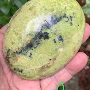 May include: A smooth, oval-shaped green stone with black and brown inclusions. The stone is held in a hand, and the surface has a slightly textured appearance. The overall colour is a pale green with darker spots.