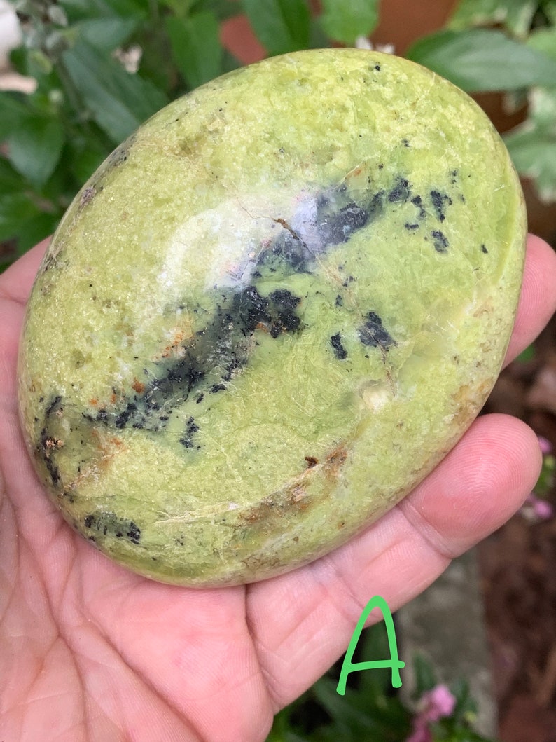 May include: A smooth, oval-shaped green stone with black and brown inclusions. The stone is held in a hand, and the surface has a slightly textured appearance. The overall colour is a pale green with darker spots.
