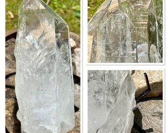 May include: A large, clear quartz crystal with a pointed tip and a rough, natural base. The crystal is sitting on a wooden surface and is illuminated by sunlight, showcasing its internal structure.