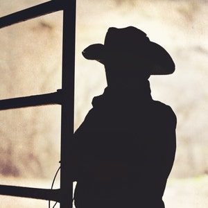 May include: Silhouette of a person wearing a cowboy hat standing in front of a wooden fence.