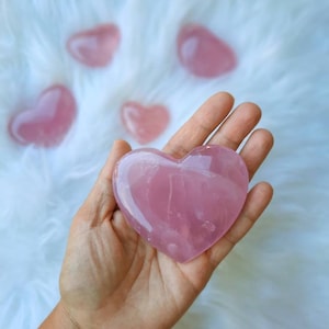 Peut inclure: Un grand cristal de quartz rose en forme de cœur de couleur rose. Le cristal est lisse et poli, avec une apparence translucide. Le cristal est tenu dans une main.