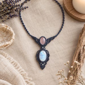 May include: A dark blue macrame necklace featuring a central oval moonstone cabochon and a smaller oval rose quartz cabochon set in intricate knotted cord work with small silver-toned bead accents along the pendant structure.