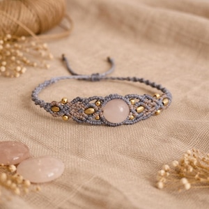 Peut inclure: Bracelet macramé fait main avec une grosse pierre de quartz rose ronde au centre. Le bracelet est gris et beige avec des perles dorées. Le bracelet est présenté sur un tissu de couleur neutre.