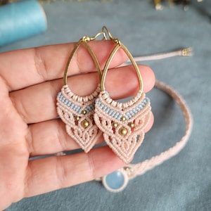 Puede incluir: Un par de pendientes de macramé rosa y azul con cuentas doradas y aros de latón. Los pendientes cuelgan de una mano.