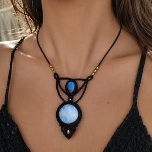 May include: A macrame necklace featuring a large circular moonstone and a smaller teardrop-shaped labradorite gemstone. The stones are set in black cord with intricate knotting, accented by small brass beads along the thin black cord necklace.
