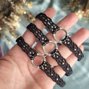 Puede incluir: Tres pulseras de macramé negras con anillos y cuentas plateadas.