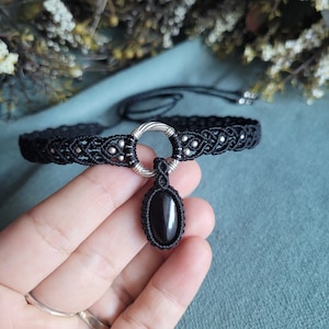 May include: Black choker necklace with a silver pendant featuring a black oval gemstone. The necklace is made of macrame cord and has a silver ring clasp.