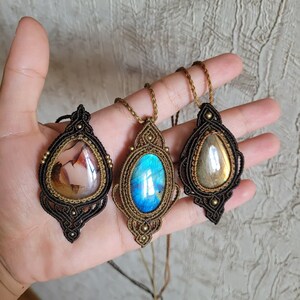 May include: Three macrame pendant necklaces featuring teardrop and oval gemstones. The left pendant has a brown and white stone in black cord, the center has a bright blue labradorite in tan cord, and the right has a gold-toned stone in black cord.