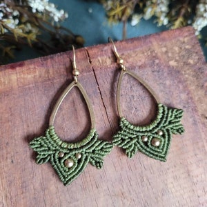 Può includere: Un paio di orecchini in macramè verde con dettagli dorati. Gli orecchini sono realizzati con un cordoncino annodato e hanno una forma a goccia con una piccola perla dorata nella parte inferiore.