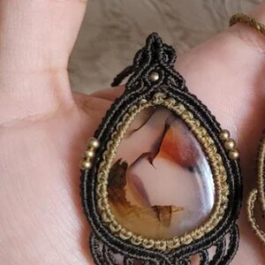 May include: A teardrop-shaped pendant featuring a polished, translucent stone with brown, orange, and cream inclusions. The stone is encased in dark brown macrame cord with intricate knotting, accented by small metallic beads along the edges.