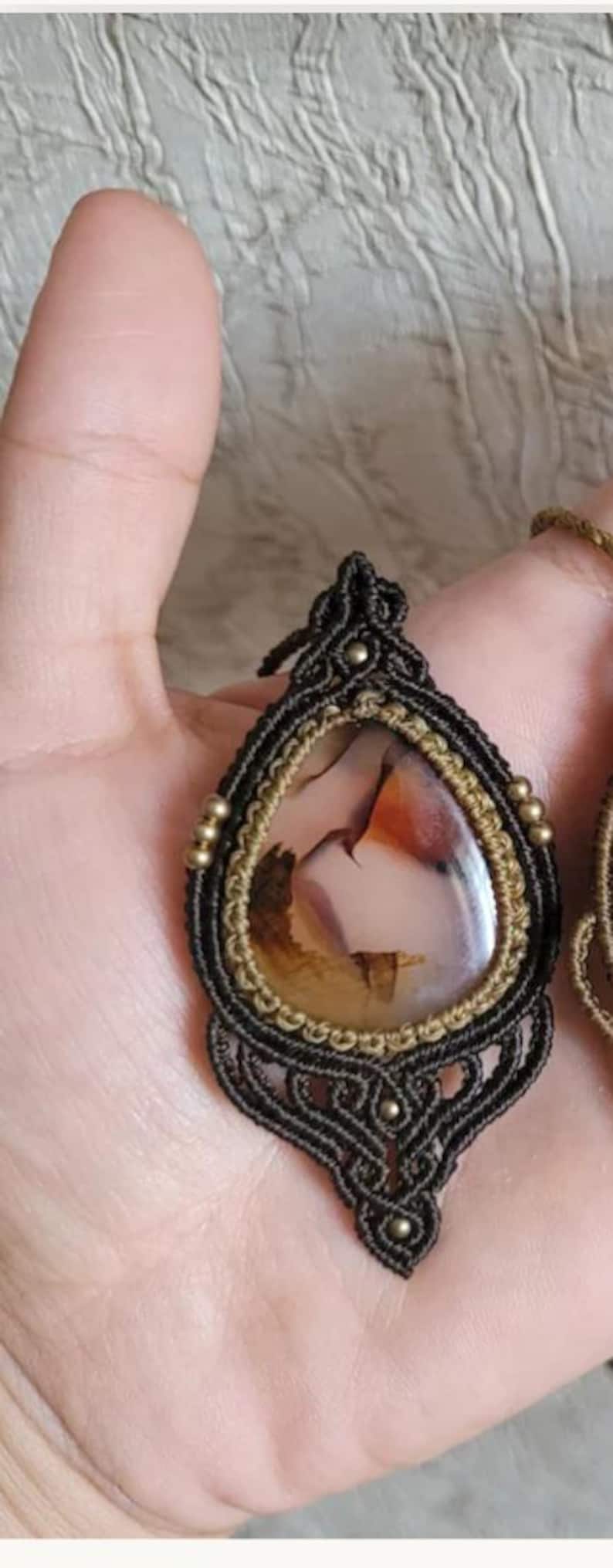 May include: A teardrop-shaped pendant featuring a polished, translucent stone with brown, orange, and cream inclusions. The stone is encased in dark brown macrame cord with intricate knotting, accented by small metallic beads along the edges.