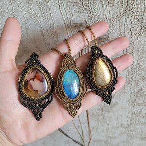 May include: Three macrame pendants featuring teardrop-shaped gemstones. The left pendant has a translucent agate stone, the center has a vibrant blue labradorite, and the right has a golden labradorite. Each is encased in intricate brown cord.