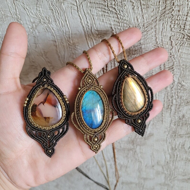 May include: Three macrame pendants featuring teardrop-shaped gemstones. The left pendant has a translucent agate stone, the center has a vibrant blue labradorite, and the right has a golden labradorite. Each is encased in intricate brown cord.