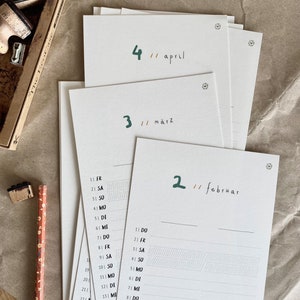 May include: A stack of white paper calendar pages with green handwritten month names and numbers. The months are April, M&auml;rz, and Februar. A pencil and a stamp set are visible in the background.