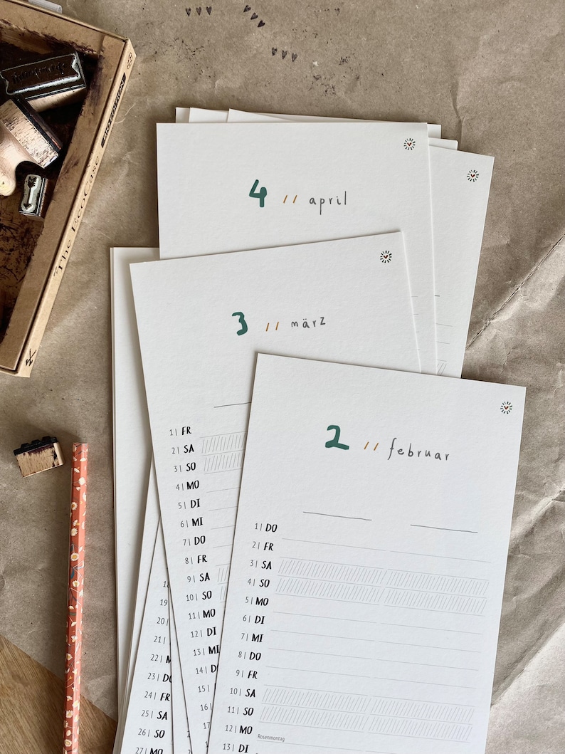 May include: A stack of white paper calendar pages with green handwritten month names and numbers. The months are April, M&auml;rz, and Februar. A pencil and a stamp set are visible in the background.