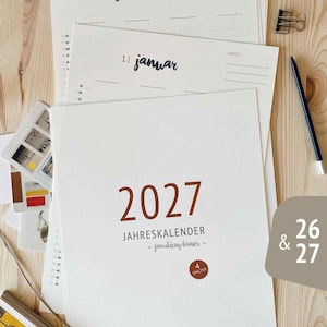 Könnte beinhalten: Ein Stapel weißer Papierkalender mit dem Jahr 2027 in brauner Schrift. Der Kalender trägt die Aufschrift "JAHRESKALENDER" und "familienplaner". Andere Seiten zeigen Daten wie "21 februar" und "11 januar". Ein Bleistift und eine Büroklammer sind sichtbar.
