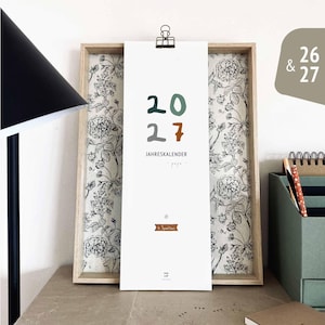 May include: A framed calendar with the year 2027 in green, brown, and gray. The calendar is held by a black clip and features the text "JAHRESKALENDER". The frame has a floral patterned background.
