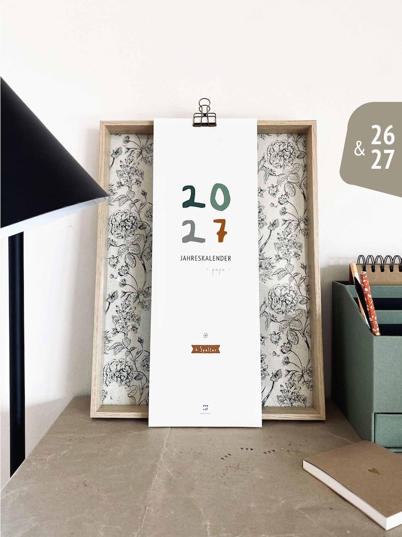 May include: A framed calendar with the year 2027 in green, brown, and gray. The calendar is held by a black clip and features the text "JAHRESKALENDER". The frame has a floral patterned background.