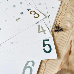 Pode incluir: V&aacute;rias folhas de calend&aacute;rio brancas com linhas de grade pretas e n&uacute;meros grandes coloridos est&atilde;o dispostas em leque. Os n&uacute;meros 3, 4, 5 e 6 est&atilde;o impressos em negrito grande com tons variados de verde e marrom. Um pequeno clipe de metal.