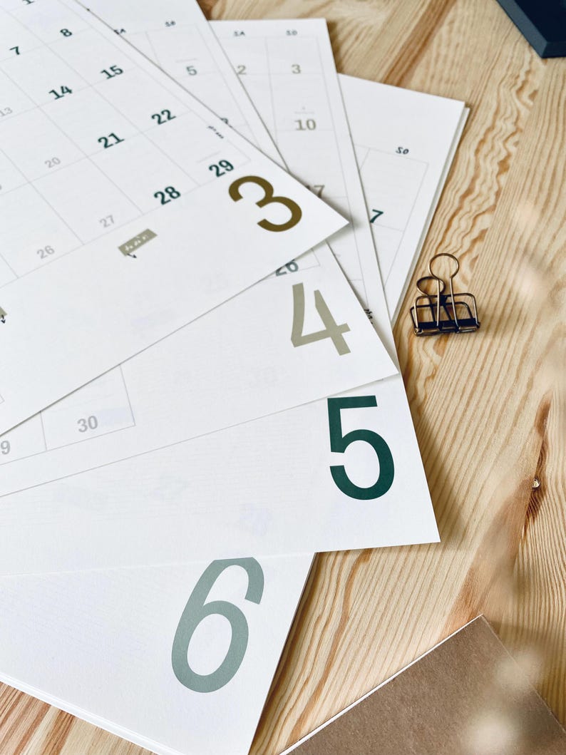 Pode incluir: V&aacute;rias folhas de calend&aacute;rio brancas com linhas de grade pretas e n&uacute;meros grandes coloridos est&atilde;o dispostas em leque. Os n&uacute;meros 3, 4, 5 e 6 est&atilde;o impressos em negrito grande com tons variados de verde e marrom. Um pequeno clipe de metal.
