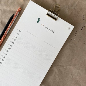 May include: A white notepad with a gold clip, featuring handwritten text and a green graphic. The date "11 august" is written on the top right. Two pencils and a small wooden block are on the left.
