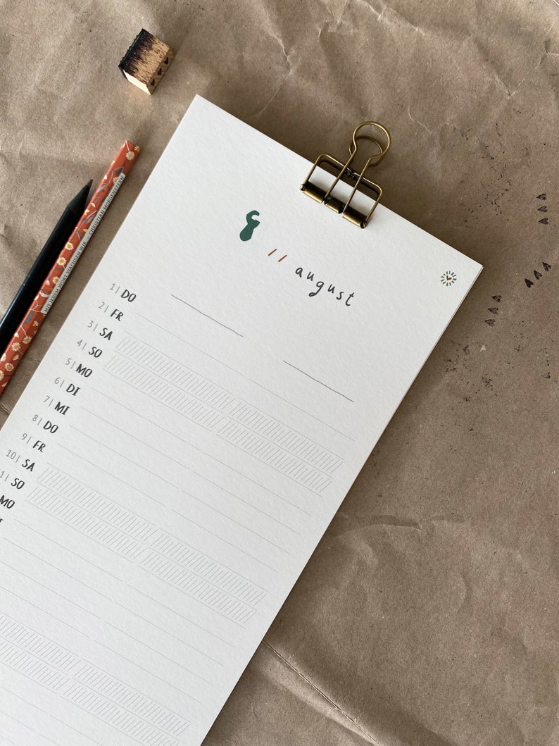 May include: A white notepad with a gold clip, featuring handwritten text and a green graphic. The date "11 august" is written on the top right. Two pencils and a small wooden block are on the left.