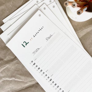 May include: A stack of white paper sheets with a calendar layout. The date "12 // dezember" is written in black ink, with the names "Frida" and "Paul" handwritten on the pages. A small white container holds several pencils with orange and white stripes.