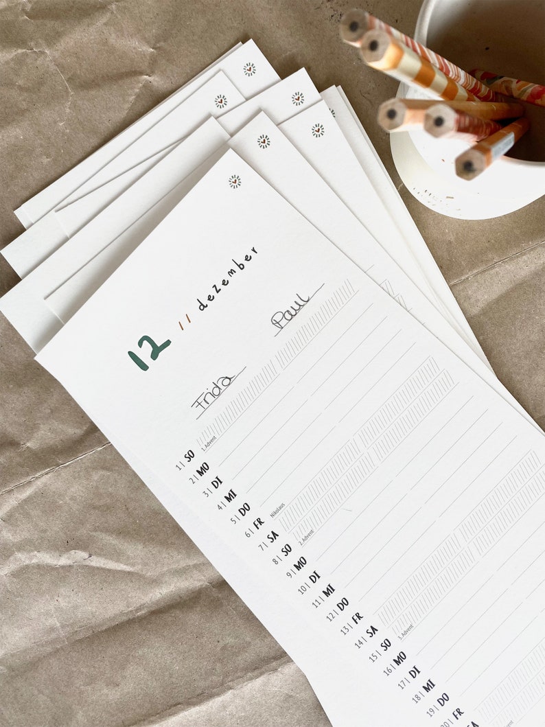 May include: A stack of white paper sheets with a calendar layout. The date "12 // dezember" is written in black ink, with the names "Frida" and "Paul" handwritten on the pages. A small white container holds several pencils with orange and white stripes.