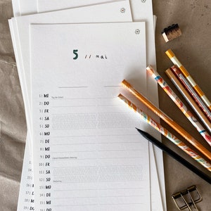 May include: A stack of white paper calendar pages with the date "5 // mai" in green and black ink. Several pencils with colorful designs and a black pencil are next to the calendar. A gold paper clip and a small stamp are also visible.