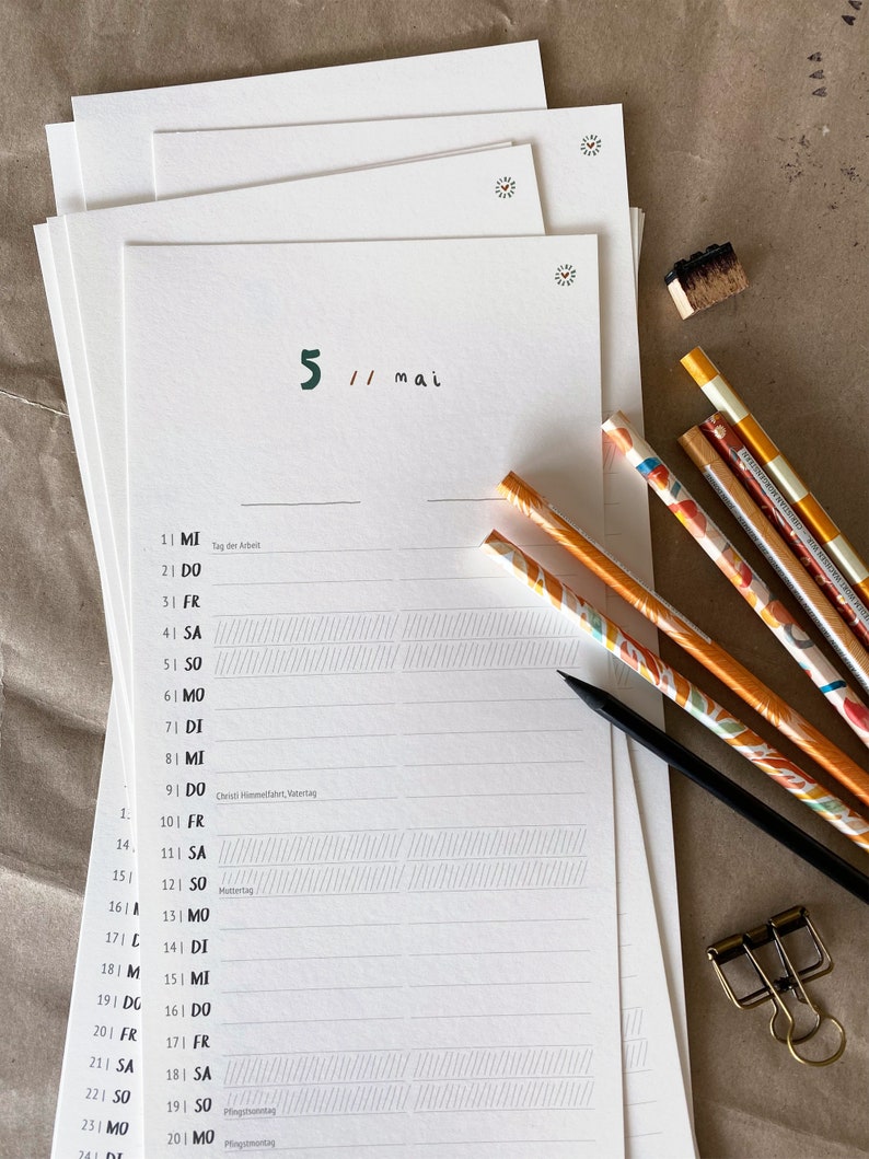 May include: A stack of white paper calendar pages with the date "5 // mai" in green and black ink. Several pencils with colorful designs and a black pencil are next to the calendar. A gold paper clip and a small stamp are also visible.