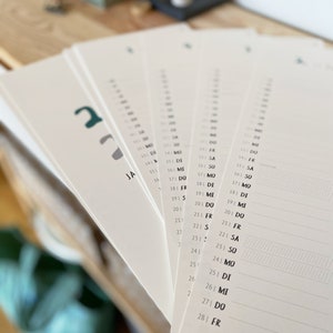 May include: A stack of white paper calendar pages fanned out, with dates and days of the week printed in black. The word "JA" is visible on one page. The calendar pages have a minimalist design.