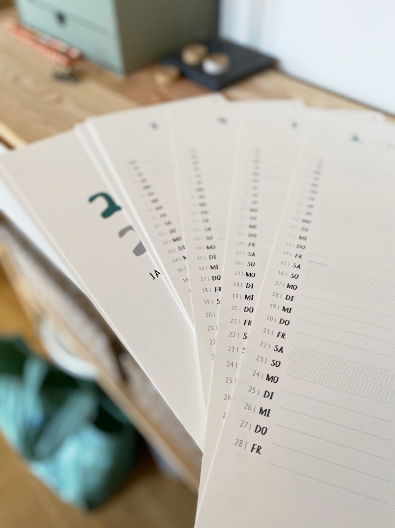 May include: A stack of white paper calendar pages fanned out, with dates and days of the week printed in black. The word "JA" is visible on one page. The calendar pages have a minimalist design.