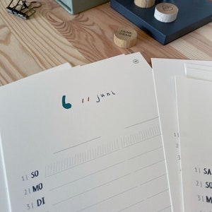 May include: A close-up of a desk setup featuring a calendar with the date "6 // juni" written in teal and black ink. Wooden circular tokens with the words "TO DO" and "DENK DRAN" are visible. Pencils, a paper clip, and a box are also present.