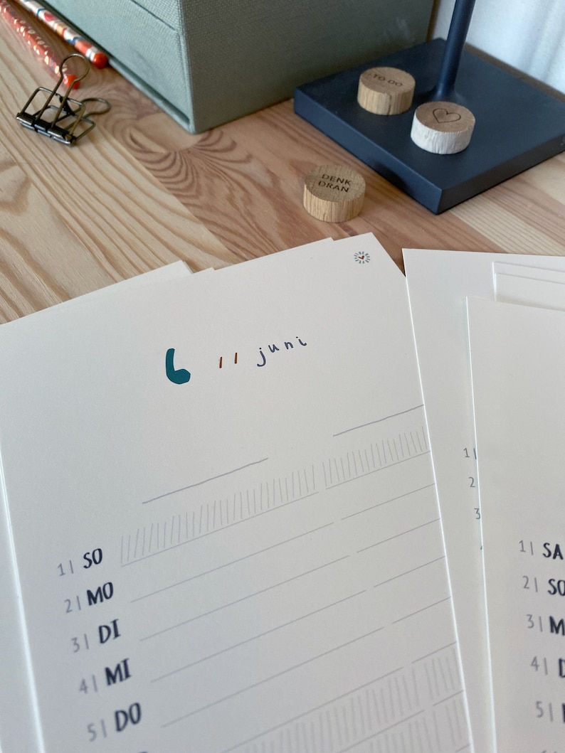 May include: A close-up of a desk setup featuring a calendar with the date "6 // juni" written in teal and black ink. Wooden circular tokens with the words "TO DO" and "DENK DRAN" are visible. Pencils, a paper clip, and a box are also present.