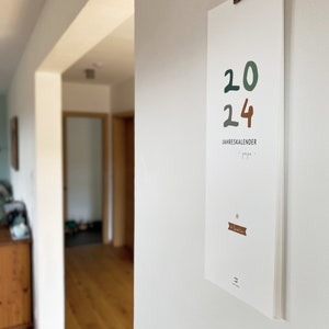 May include: A white 2024 calendar hanging on a wall. The numbers "20" are in green, and "24" is in brown. The text "JAHRESKALENDER" is below the numbers. The calendar is held by a metal clip.