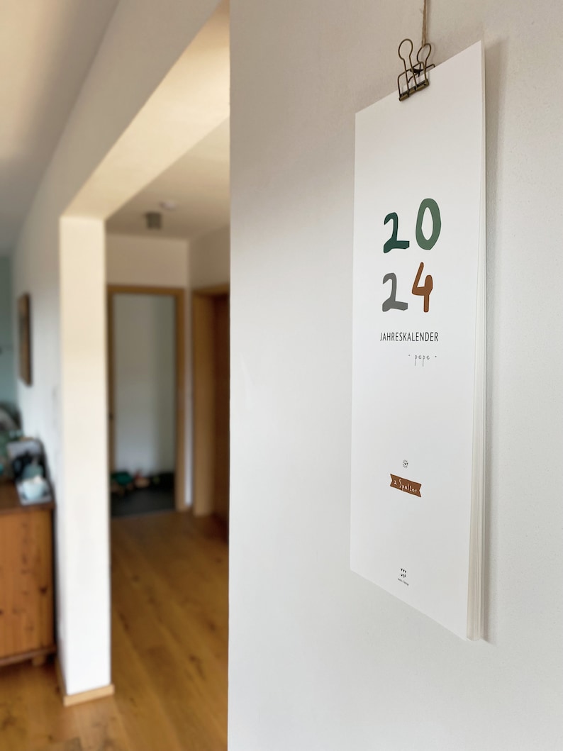 May include: A white 2024 calendar hanging on a wall. The numbers "20" are in green, and "24" is in brown. The text "JAHRESKALENDER" is below the numbers. The calendar is held by a metal clip.