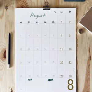 Pode incluir: Um calend&aacute;rio de parede branco para o m&ecirc;s de agosto com um clipe de metal preto no topo. O calend&aacute;rio apresenta um layout de grade com os dias da semana marcados como MO, DI, MI, DO, FR, SA, SO e um grande n&uacute;mero 8 dourado no canto inferior direito.